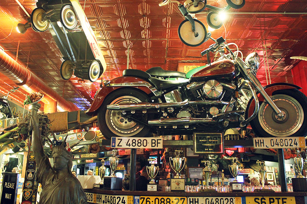 Интерьер бара напоминает музей: всюду мотоциклы и машины, автомобильные номера, значки и трофеи. К факелу статуи Свободы прилепили тарелку с&nbsp;куриными крылышками — это тоже символ Америки