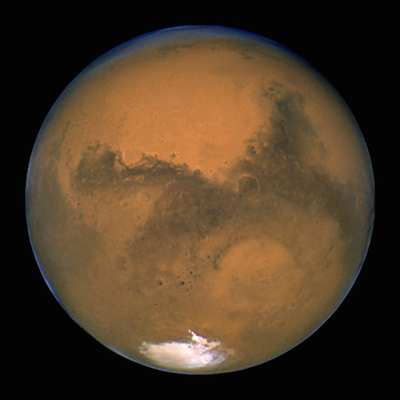 A photo of one full lit side of Mars, showing the reddish brown color of its surface and a white spot on the southern side.