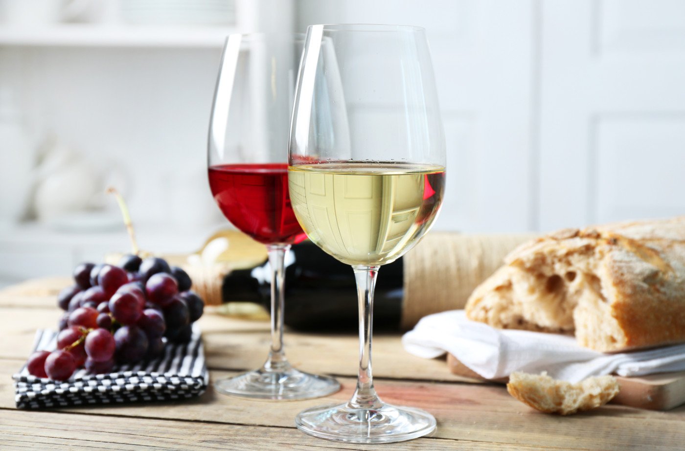 На фото красное и белое вино в бокалах на фоне свежевыпеченного хлеба, бутылки вина и сочного винограда